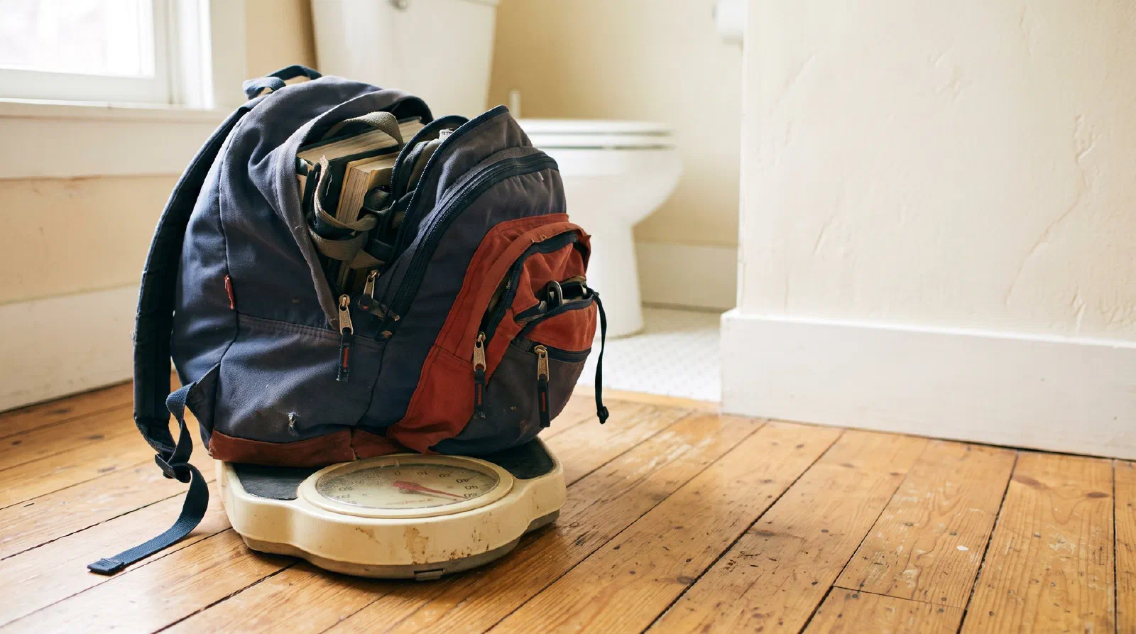 A heavy overstuffed school backpack sitting on a bathroom scale on a wooden floor